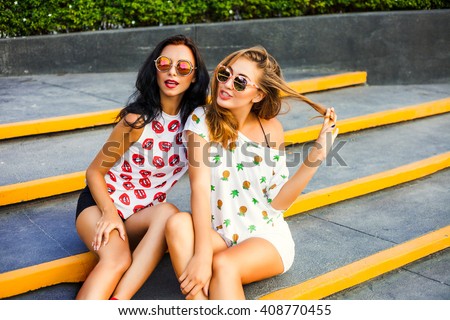 Two trendy hipster cool tanned bold girl, and ride in the shopping cart. Dressed in colorful T-shirts with his lips and pineapple, shorts, sunglasses.  Shopping.