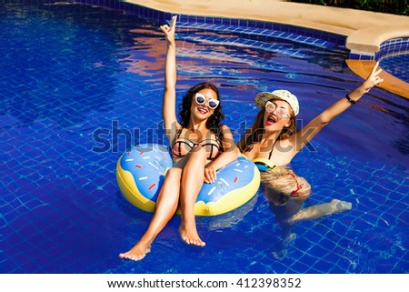 Two beautiful sexy fun cool hipster girls swim in the pool on an inflatable circle, donut. Bright swimsuit, cap, the girls laugh, in sunglasses blonde and brunette, velvety skin, red lips, pool party.