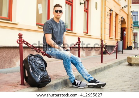 Young handsome man sitting on the street. Dressed in a T-shirt, jeans, fashion sneakers, with a backpack in the form of a lion, sunglasses, Hipster style guy