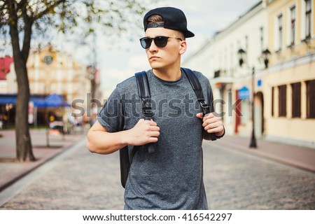Young free beautiful fashionable hipster guy walking down the street, through the park, wearing a cap with a backpack, t-shirt, sunglasses, urban style, sport casual