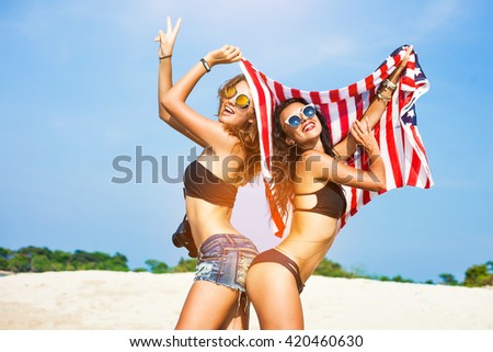 Two beautiful tanned fun hipster girls on the beach, blonde and brunette standing back to back and holding an American flag, old style, laugh, smile relax on a tropical island sexy bikini denim shorts