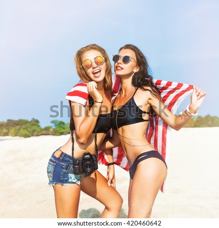 Two cool trendy hipster girls at the beach enjoying vacation on a tropical island, perfect tanned body, sexy stylish casual wear, bikinis, blonde and brunette with the camera, woman photographer