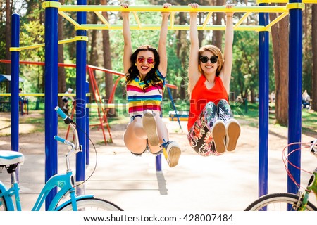 Two beautiful happy funny hipster girls, best friends, hang on the crossbar on the horizontal bar on the playground, laughing, play the fool, fun watching, a great sunny summer day, Lifestyle
