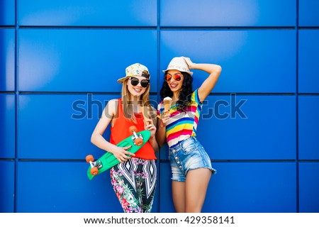 Two teen girl friends having fun together with skate board. Outdoors, urban lifestyle. eat ice cream, wearing denim shorts, colorful leggings, bright T-shirts, rainbow, sunglasses, cap, hat, hipster