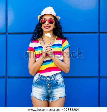 Beautiful cute funny amazing young hipster teen girl eating ice cream cone, laughs happy, bright casual wear, denim shorts, striped, colored T-shirt, rainbow, blue background, urban style, sunglasses