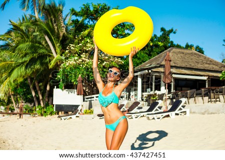 Beautiful fashionable sexy cute happy girl running on the beach with a yellow rubber ring in the hands on the background of the hotel, trendy turquoise swimsuit, sunglasses, hairstyle with horns