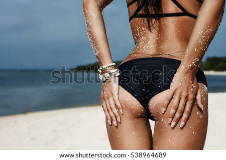 Slim luxury girl in a black bikini on the beach. Photos from behind. Perfect tanned body, sexy ass, perfect figure. Rest on a tropical island. Golden bracelets Photo in low key. Vogue Style