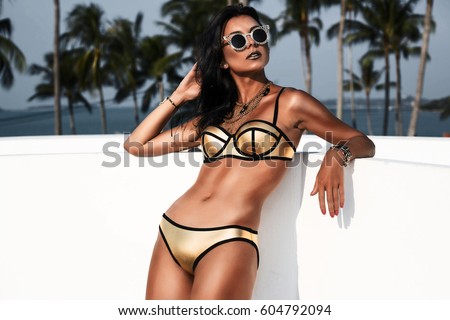 Fashion portrait of a beautiful young sexy woman. Dressed in a gold swimsuit with gold accessories, bracelets and necklaces, tanned skin,l body, against a backdrop of palm trees, a photo in a low key
