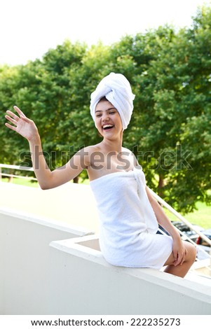 Young happy girl in a towel after a shower laughing and waving on the balcony on a summer day