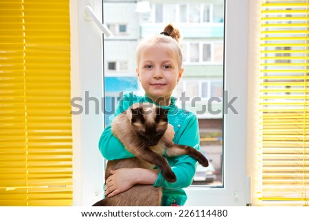 Little girl with a cat in hands sitting at the window with yellow blinds in the room