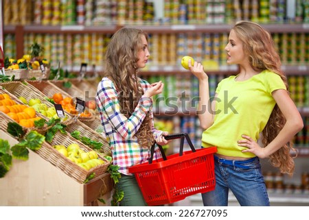 Two girls at the grocery store with a red basket in hand pick fruits