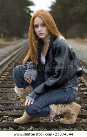 Pretty red haired girl in a black leather jacket crouching on train tracks