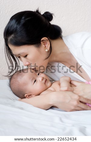 Asian mother and baby in bed; mother kissing baby\'s forehead