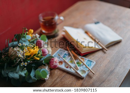 Artist\'s desktop with watercolors, palette, sketchbook. Still life. Wooden table with a spring bouquet of flowers, painting supplies and a mug of tea.