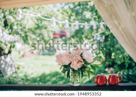 Still life of tea accessories and flowers. A pair of bright mugs of tea, a bouquet of peonies on the veranda on a background of a green garden on a bright day