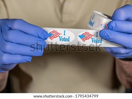 Hands in medical gloves holding roll of I Voted buttons with USA Flag ready for in person voters in Presidential election