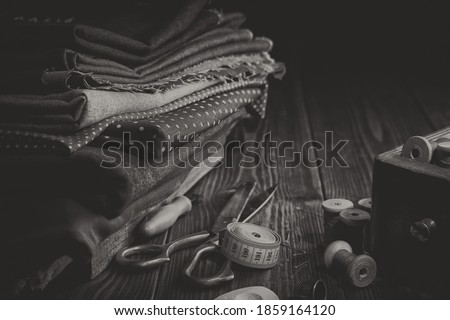 Stack of fabrics for sewing, scissors, measuring tape, spools of thread, wooden box of sewing accessories. Black and white styled photo.
