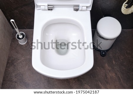 Clean toilet bowl with drain hole, top view. Toilet brush and trash can on black tiles