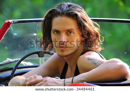 long hair male model sitting in a red convertible classic sport car