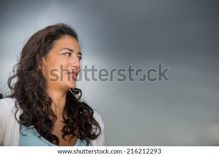 Beautiful brown young woman (20s) on profile looking to the right with smiling. Head and shoulders on cloudscape background. Right copy space.
