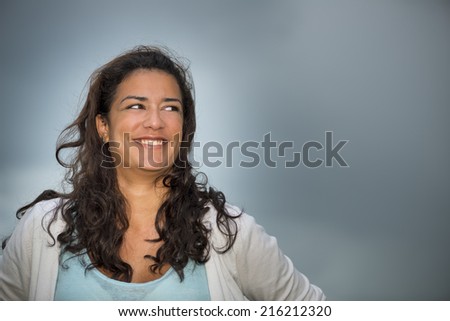 Beautiful brown young woman (20s) looking to the right with smiling. Head and shoulders on cloudscape background. Right copy space.