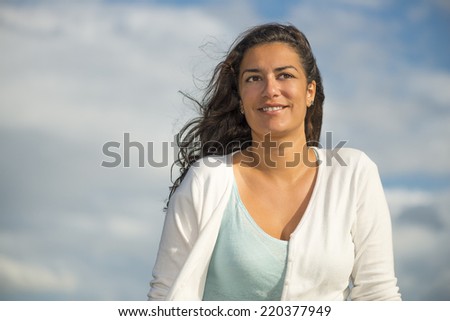 Head and shoulders of beautiful young woman smiling. She is looking off camera by day wind with long brown hairs. Cloudscape background with blue sky