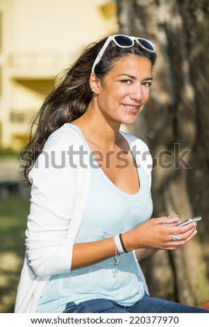 Portrait outdoors of an attractive young woman sitting with sun glasses on the top of her head. She is smiling and looking at camera with her smartphone. Head and shoulders.