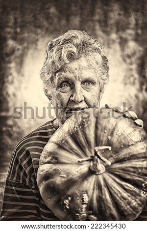 Halloween theme. Portrait of an old woman holding a big ripe pumpkin freshly harvest in autumn. Studio shoot and sepia toned image.