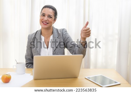 Young woman well-dressed smiling with brown hair back sitting at her desk giving a single thumbs up to the camera. Business portrait.
