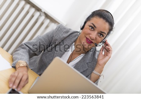 Customer service representative in action sitting with headset on at her wood desk behind laptop computer. She is smiling and looking at camera.