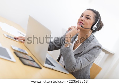 Smiling well-dressed saleswoman with headset on in office behind her desk and laptop computer. Copy space