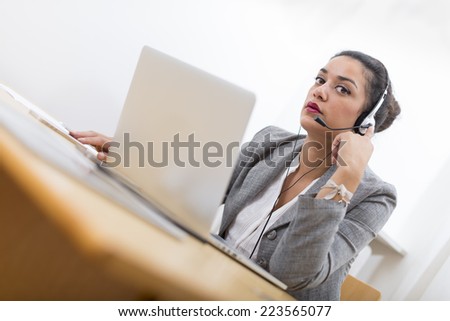 Serious well-dressed saleswoman with headset on in office behind her desk and laptop computer. Copy space