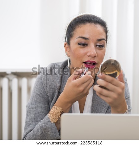 Portrait of a beautiful well-dressed businesswoman applying lipstick with little mirror in office. Action before a meeting or before to leave the office.