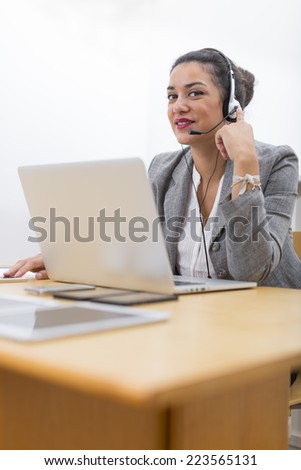 Attractive young woman in office enjoying working with headset on. She is smiling behind her laptop computer on wood desk.