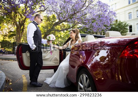 Happy groom helping his bride out of the cabriolet red car; newly wedded standing near wedding car.