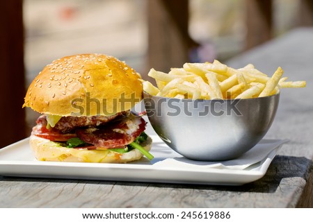 Tasty burger with melted cheese and a thick succulent ground beef patty garnished with lettuce, tomato, onion and rocket on a sesame bun standing on a table with french fries