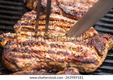 Beef steaks being prepared on grill with BBQ fork and knife with pepper, spices and seasoning with spatula