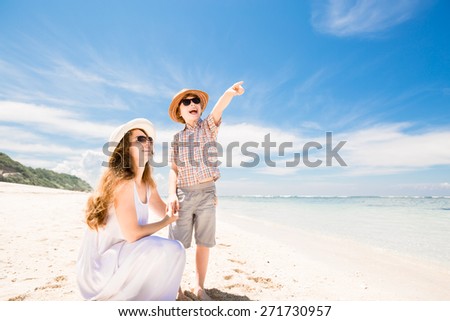Happy beautiful mother and son walking along the beach in a sunny day with deep blue sky over the ocean on background