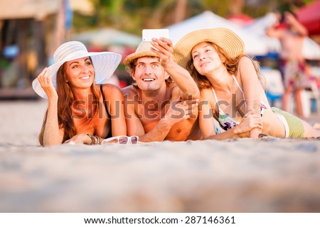 Group of happy young people in bathing suits lying on the beach with wite sand and having fun in sunset sun and taking selfie