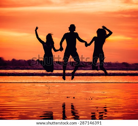 Silhouettes of happy young friends jumping on the beach  on sunset with beautiful red sky on background