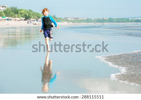 Young boy in swimming shorts and rushwest runs along Bali beach near sunset with reflection in the water