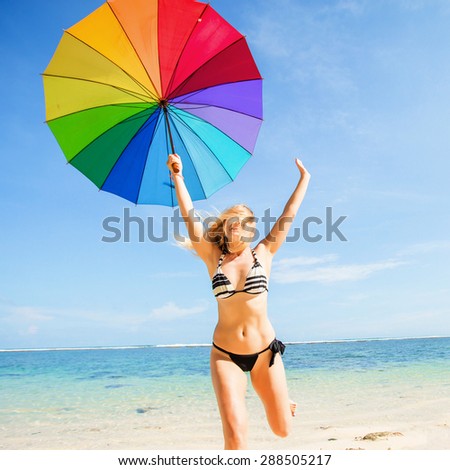 Young skinny girl in blue shorts with colourful rainbow umbrella jumps on the beach with clear blue sky and ocean on background