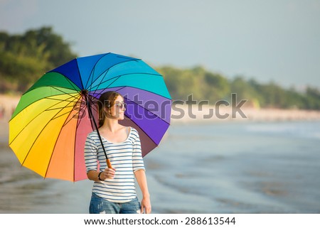 Young caucasian woman wearing white sunglasses with colourful rainbow umbrella looking at ocean on the Jimbaran beach on Bali before sunset. Travel, holidays, vacation, healthy lifestyle, tranquility
