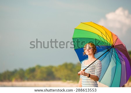 Young caucasian woman wearing white sunglasses with colourful rainbow umbrella looking at ocean on the Jimbaran beach on Bali before sunset. Travel, holidays, vacation, healthy lifestyle, tranquility