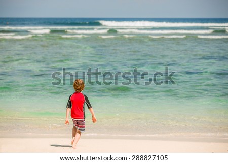 Cute 7 years old boy in red rash vest swimming suit and shorts having fun on tropical Bali beach with white sand and green ocean