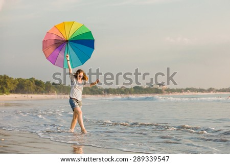 Cheerful caucasian young woman with rainbow umbrella having fun on the Jimbaran beach on Bali before sunset with beautiful ocean and blue sky on background. Travel, holidays, vacation, healthy