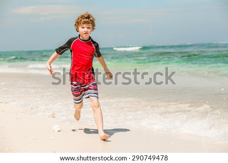 Cute young boy in red rushwest swimming suit and shorts having fun on summer holidays on tropical Bali beach with white sand and green ocean
