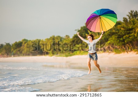 Cheerful caucasian young woman with rainbow umbrella having fun on the Jimbaran beach on Bali before sunset with beautiful ocean and blue sky on background. Travel, holidays, vacation