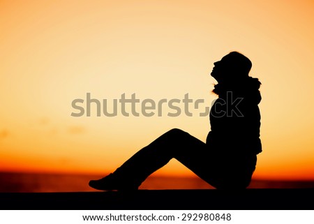 Silhouette of a woman in coat sitting on the edge with beautiful red sunset on background. Peace, cerenty, lifestyle concept