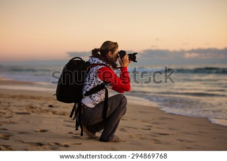 Young woman photographer with backpack taking pictures with SLR camera at sunset on the winter beach.